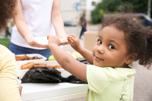 Child in a yellow shirt receives food from an adult at an outdoor table, with plates of food visible.